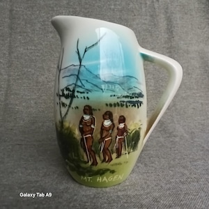 May include: A cream-coloured ceramic pitcher featuring a hand-painted landscape scene with three figures standing in the foreground. The artwork depicts mountains and foliage in blue and green tones. The text MT. HAGEN is printed at the base.