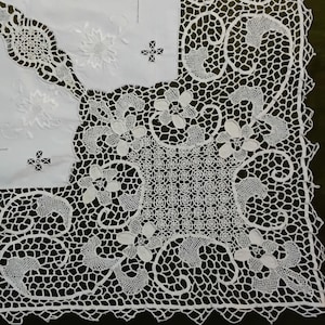 May include: A close-up view of a white lace tablecloth with detailed floral and geometric patterns. The design includes openwork and embroidered elements, creating a delicate texture. The tablecloth is square in shape.