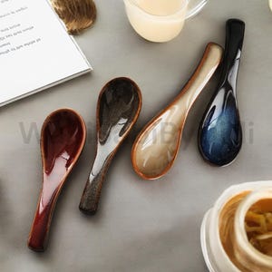Könnte beinhalten: Fünf Keramiklöffel in verschiedenen Farben: Braun, Dunkelbraun, Beige, Blau und Rot. Die Löffel sind in einer Reihe auf einer grauen Oberfläche angeordnet.