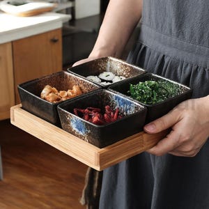 May include: A set of four black and blue ceramic bowls with a wooden tray. The bowls are filled with different foods, including seaweed, red and white fish, and a brown sauce.