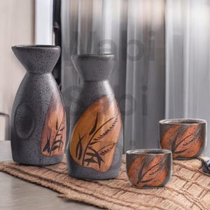 May include: A set of four ceramic sake bottles and cups with a black and brown speckled glaze and a leaf design. The bottles and cups are arranged on a woven mat.