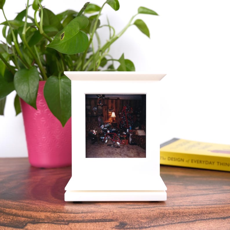 May include: A white photo frame with a picture of a Christmas scene. The frame is on a wooden surface, with a pink pot of greenery to the left. A yellow book is visible to the right.