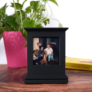 May include: A black wooden picture frame with a photograph of two women. The frame is rectangular with a decorative top and base. A pink flower pot with a green plant is in the background.