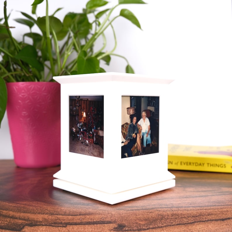 May include: A white photo display with two visible picture frames. The display is square with a layered design. A pink potted plant and a book are in the background. The photos inside the frames show people and a Christmas tree.