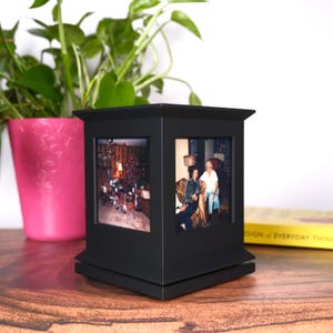 May include: A black, four-sided photo cube with a decorative top and base. The cube displays two framed photos. A pink pot with a green plant is in the background, along with a yellow book. The cube sits on a wooden surface.