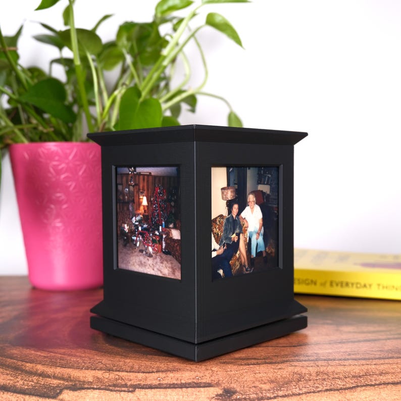 May include: A black, four-sided photo cube with a decorative top and base. The cube displays two framed photos. A pink pot with a green plant is in the background, along with a yellow book. The cube sits on a wooden surface.