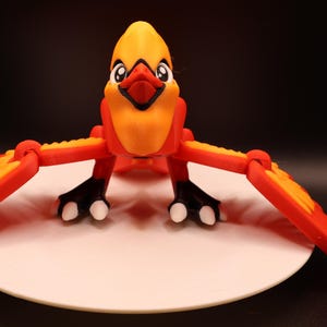 以下が含まれることがあります： 翼を広げた、鮮やかな漫画風の鳥の置物。鳥は黄色い頭と胸、赤い嘴、赤と黄色の翼が特徴です。足は白いつま先のある黒色で、白い円形の台座の上に立っています。