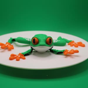 May include: A green and orange frog figurine with white spots. The frog is posed on its belly with its legs outstretched. The figurine is on a white plate.