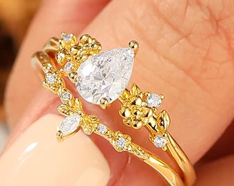 May include: A gold-colored ring set featuring a pear-shaped, clear gemstone and smaller accent stones. The rings have a floral design with delicate details. The rings are on a finger with a light-colored nail polish.