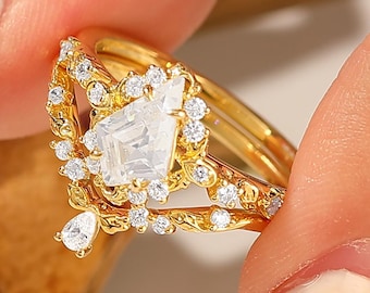 May include: A gold-toned ring with a large, square, clear gemstone at the center, surrounded by smaller, clear stones. The ring has a delicate, ornate design with a teardrop-shaped stone hanging from the bottom. The ring is held by a person.