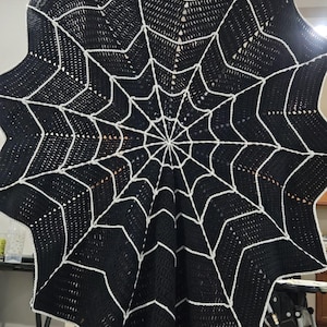 Puede incluir: Manta de ganchillo de araña negra y blanca. La manta está hecha con hilo negro y tiene un diseño de telaraña de ganchillo blanco.