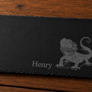 May include: A rectangular black slate serving board with a rough, uneven edge. The surface features a laser-engraved illustration of a bearded dragon lizard on the right side and the name Henry engraved in a serif font on the lower left side.