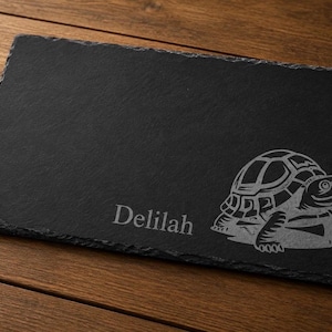 May include: A rectangular black slate serving board with a textured surface. The board features a detailed engraved turtle design and the name "Delilah" in a stylish font. The board rests on a wooden surface.