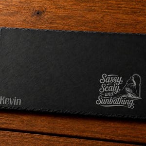 May include: A rectangular black slate serving board with a rough edge. The board has the name "Kevin" engraved on the left side. The phrase "Sassy, Scaly, and Sunbathing" is engraved on the right side, along with a graphic of a frog under a lamp.