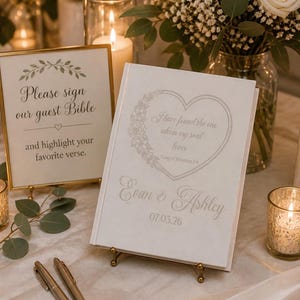 May include: A white guest Bible book featuring a heart-shaped floral design, the text I have found the one whom my soul loves, Song of Solomon 3:4, the names Evan & Ashley, and the date 07.03.26, displayed on a gold stand with two pens.
