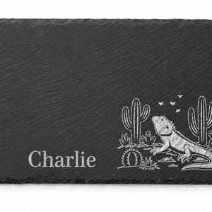 May include: A rectangular black slate board with rough, uneven edges. The surface features an engraved white graphic of a bearded dragon lizard surrounded by cacti and small birds, with the name Charlie engraved in white text on the left side.