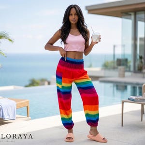 May include: A light pink sleeveless crop top paired with loose-fitting, high-waisted pants featuring a vibrant horizontal rainbow stripe pattern, a wide elasticized waistband, and cuffed ankles, worn with simple light peach-colored slide sandals.
