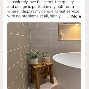 May include: A small rectangular wooden stool with four legs, supporting a white ribbed planter with green foliage and a glass reed diffuser with black sticks. The stool is positioned next to a white bathtub against a tiled bathroom wall.