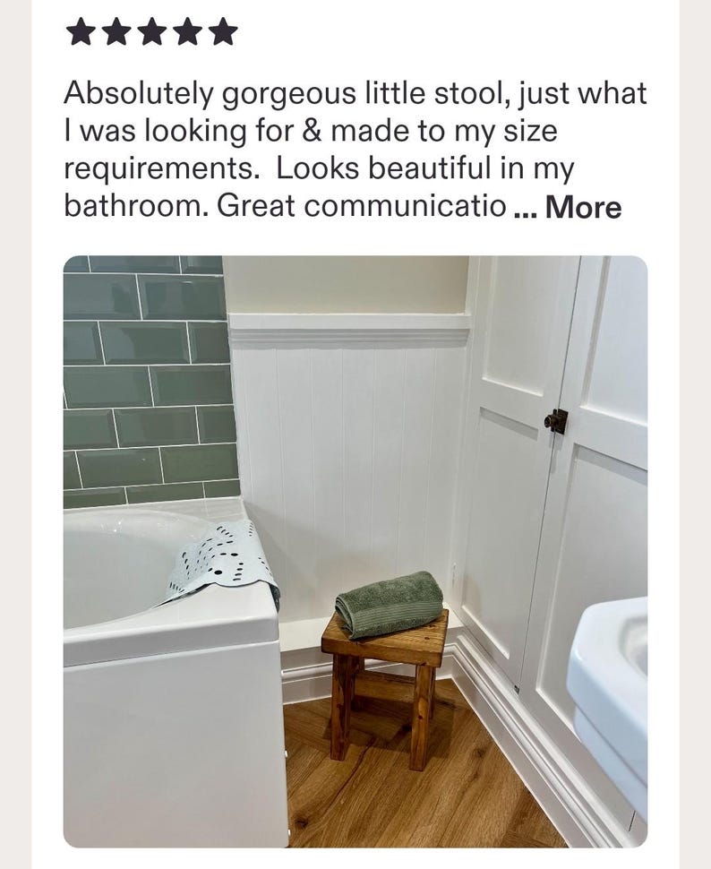 May include: A small, rectangular wooden stool with a dark brown finish and four straight legs. A folded, sage green textured towel rests on the flat top surface of the stool. The stool is positioned on a light brown wood floor in a bathroom setting.