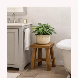May include: A square wooden stool with four legs and a cross-brace support, topped with a green leafy plant in a textured cream-colored woven basket. The text "Perfect for Bathrooms too" is displayed above the stool in a dark brown font.