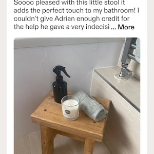 May include: A small, rectangular wooden stool with a natural finish, featuring a black plastic spray bottle, a clear glass candle with a white label, and a folded grey towel placed on its flat top surface in a bathroom setting.