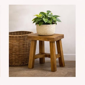 May include: A small, four-legged wooden stool with a natural brown finish, supporting a white woven basket planter containing a lush green leafy plant, positioned next to a large, tall, brown woven wicker basket with a cylindrical shape.