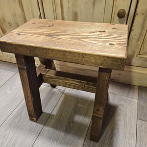 May include: A rectangular wooden stool featuring a dark brown stained finish with visible wood grain patterns. The stool is constructed with four sturdy legs connected by horizontal support beams, showcasing a simple, functional design.