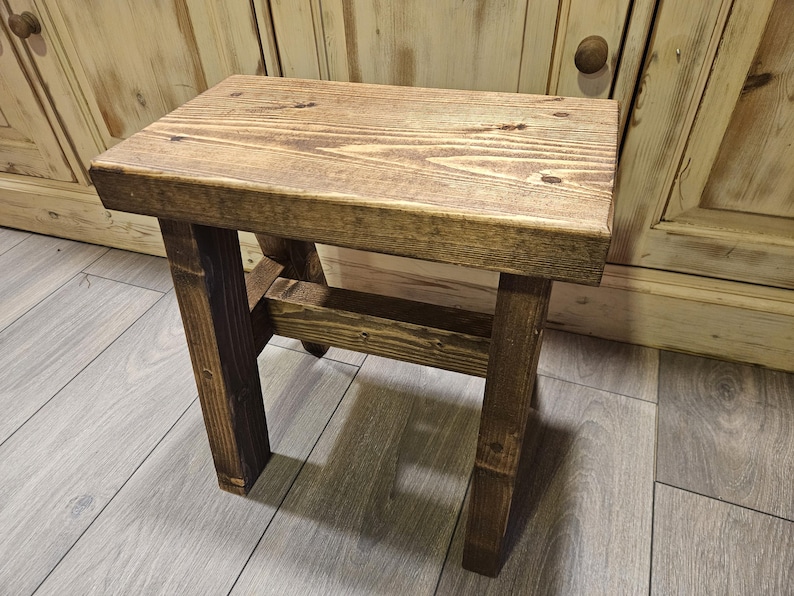 May include: A rectangular wooden stool featuring a dark brown stained finish with visible wood grain patterns. The stool is constructed with four sturdy legs connected by horizontal support beams, showcasing a simple, functional design.