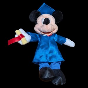 May include: A plush Mickey Mouse figure dressed in a blue graduation gown and cap, holding a rolled diploma with a red ribbon. The figure has black ears, a smiling face, and black shoes. The background is black.
