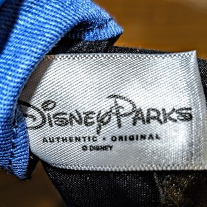 May include: A close-up of a white tag with the text "Disney Parks" in black script. Below the logo, the words "AUTHENTIC &bull; ORIGINAL" are printed. The tag is attached to a blue and black fabric.
