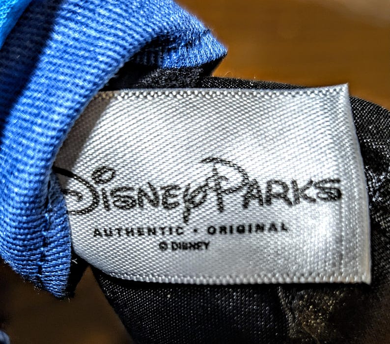 May include: A close-up of a white tag with the text "Disney Parks" in black script. Below the logo, the words "AUTHENTIC &bull; ORIGINAL" are printed. The tag is attached to a blue and black fabric.