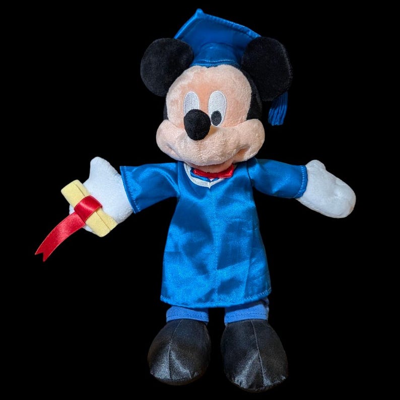 May include: A plush Mickey Mouse doll dressed in a blue graduation gown and cap, holding a diploma with a red ribbon. The doll has black ears, a black nose, and a red bow tie. The background is black.