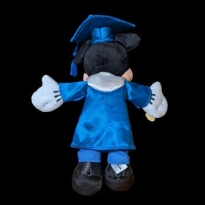 May include: A plush Mickey Mouse figure, viewed from the back, wearing a blue graduation cap and gown. The figure has black ears, a white collar, white gloves, and black shoes. The graduation attire is a vibrant blue color.