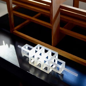 Puede incluir: Un modelo de edificio de plástico blanco con una estructura de rejilla. El modelo está sentado sobre una superficie negra y está iluminado desde arriba, creando un efecto brillante, casi translúcido.