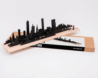 May include: A black New York City skyline silhouette model mounted on a light-colored wooden base. The model rests on a matching cardboard packaging box featuring the text NEW YORK and the brand name Be-Malevich printed on the side.