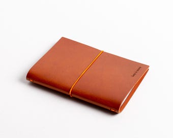 Puede incluir: Un cuaderno de cuero marrón con cierre de banda elástica naranja. El cuaderno tiene el texto "Less is more" grabado en relieve en la portada.