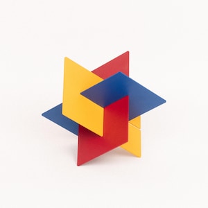 Könnte beinhalten: Eine geometrische Skulptur aus ineinandergreifenden, eckigen Formen in den Grundfarben Rot, Gelb und Blau. Das Design weist eine sternartige Form mit mehreren Ebenen und Kanten auf, vor einem weißen Hintergrund.