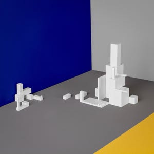 Può includere: Forme geometriche bianche disposte a forma di torre su una superficie grigia con uno sfondo blu e giallo.