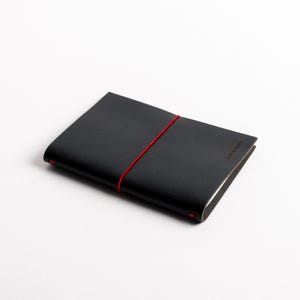 Puede incluir: Un cuaderno de cuero negro con cierre de banda elástica roja. El cuaderno tiene las palabras "Less is more" impresas en la esquina inferior derecha. El cuaderno está cerrado y plano sobre una superficie blanca.
