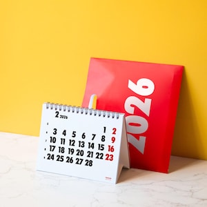 May include: A white desk calendar for February 2026 with black numbers and red highlighted dates. A red bag with the year 2026 in large white text leans against a yellow background. The calendar is spiral-bound and stands upright.