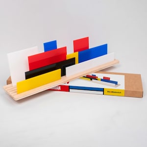 Può includere: Un vassoio di legno contiene pezzi rettangolari colorati in acrilico rosso, blu, giallo, nero e bianco. Il vassoio è posto sopra una scatola con un design simile e la scritta "BeaMalevich". Il design è ispirato all'opera di Piet Mondrian.