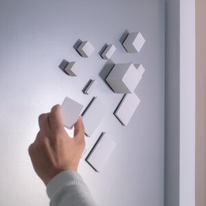May include: A hand reaches for a white cube, one of many white cubes arranged in a geometric pattern on a white wall.