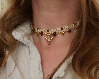 May include: A cream-colored macrame choker necklace featuring a central gold-toned bull skull charm, accented with small dark brown beads and metallic gold-toned beads woven into a scalloped pattern along the cord.