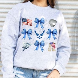 May include: A light grey sweatshirt with patriotic watercolour designs. The design includes an American flag, blue bows, an Air Force emblem, a dog tag, a fighter jet, and combat boots with an American flag.