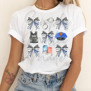 May include: White t-shirt with a blue and black watercolor design featuring police badges, handcuffs, and bows. The text "Our Police Matter" is written in a gray font.