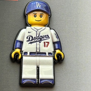 May include: A Los Angeles Dodgers baseball player minifigure pin featuring a yellow head, a blue cap with the LA logo, and a white jersey with the Dodgers script, the number 17 in red, and blue sleeves. The figure has white pants with blue trim.
