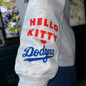 May include: A light gray long-sleeve sweatshirt featuring a graphic on the sleeve with the text HELLO KITTY in red, a small red heart, and the word Dodgers in blue script. A partial graphic of Hello Kitty is visible on the upper chest area.