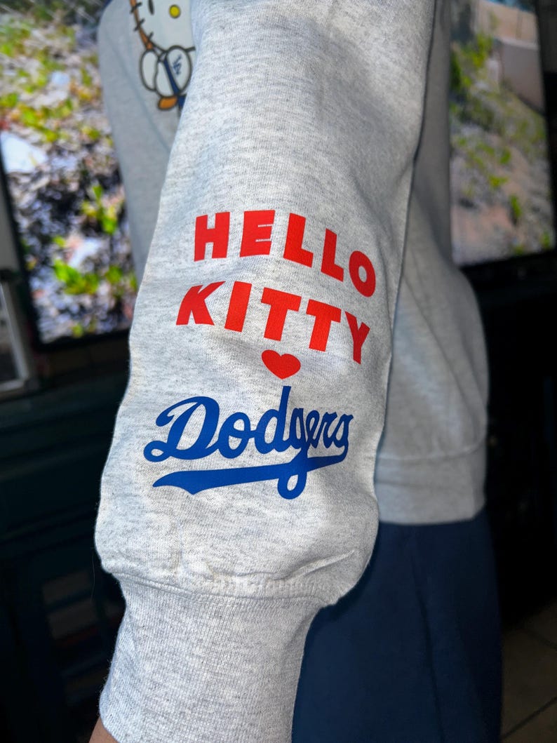 May include: A light gray long-sleeve sweatshirt featuring a graphic on the sleeve with the text HELLO KITTY in red, a small red heart, and the word Dodgers in blue script. A partial graphic of Hello Kitty is visible on the upper chest area.