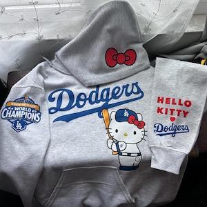 May include: A light gray hooded sweatshirt featuring the blue Dodgers logo, a graphic of Hello Kitty holding a baseball bat, and a red bow on the hood. The sleeve displays a 24 World 25 Champions patch, while the other sleeve reads HELLO KITTY Dodgers.