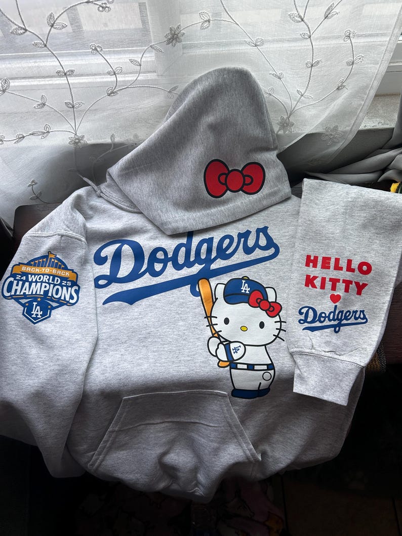 May include: A light gray hooded sweatshirt featuring the blue Dodgers logo, a graphic of Hello Kitty holding a baseball bat, and a red bow on the hood. The sleeve displays a 24 World 25 Champions patch, while the other sleeve reads HELLO KITTY Dodgers.